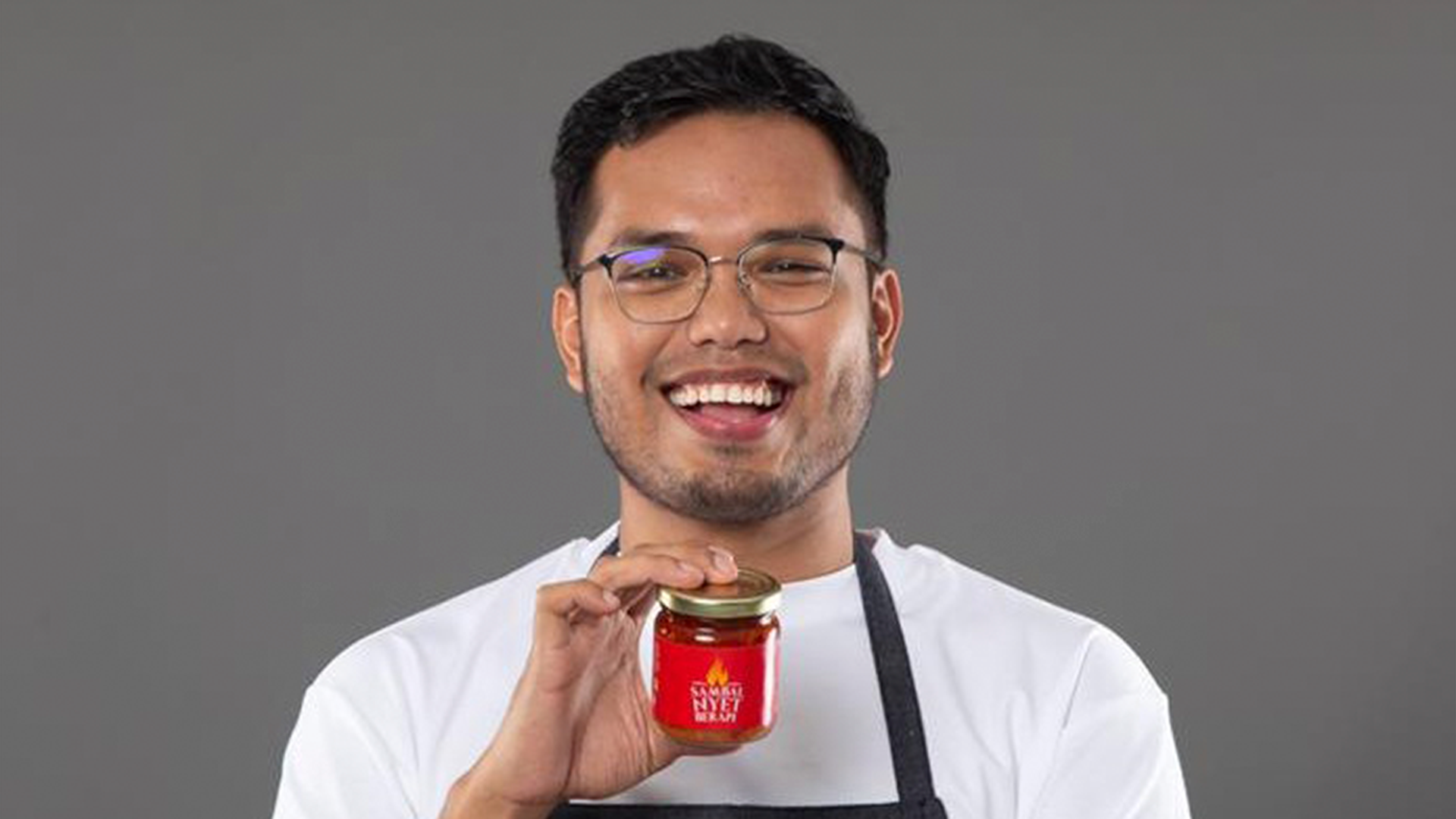a man wearing spectacle,white shirt, black apron, holding a product