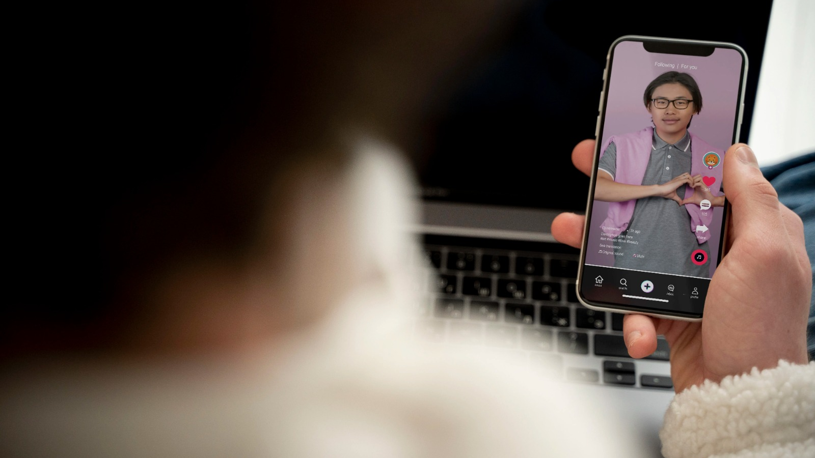 Person holding a smartphone watching a TikTok video of a young man making a heart gesture, with a laptop in the background.