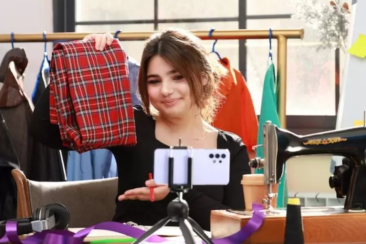 A young female content creator smiles at her smartphone, which is mounted on a tripod, as she holds up a piece of red plaid fabric. She appears to be in a craft studio, with a sewing machine, ribbons, and a rack of clothes visible in the background.