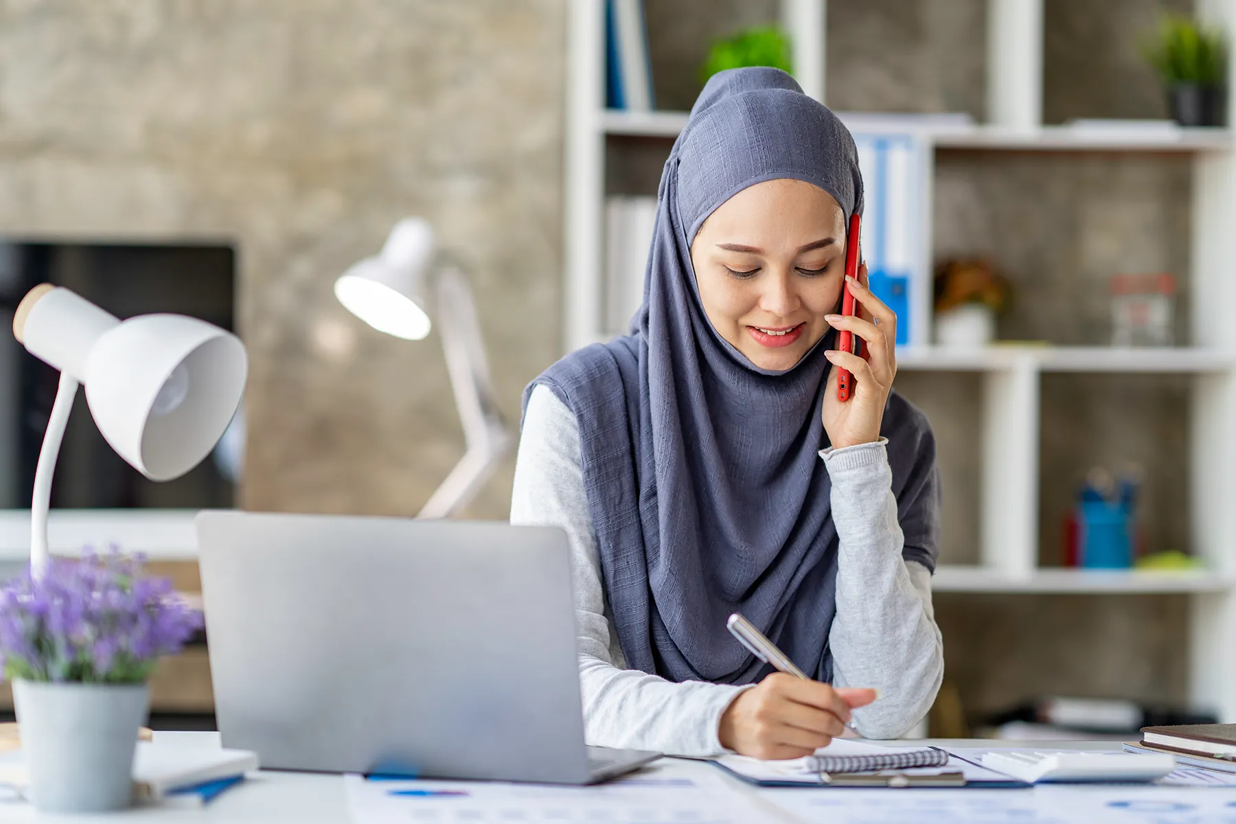 Muslim woman working at desk while talking on the phone and taking notes