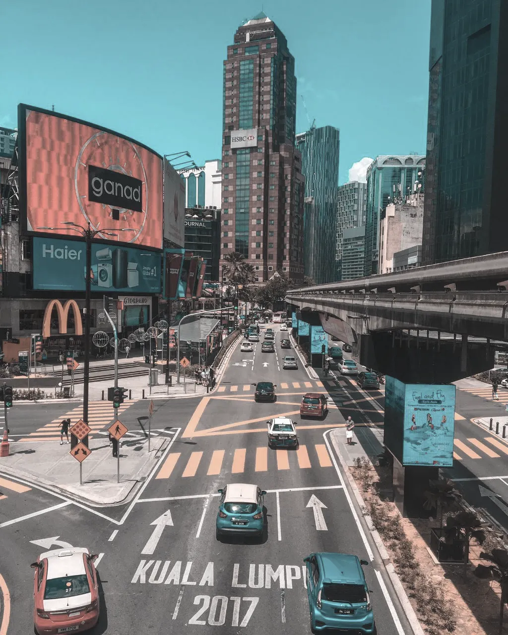 Urban view of Kuala Lumpur featuring the monorail track, large billboards, and vehicles at a major intersection.