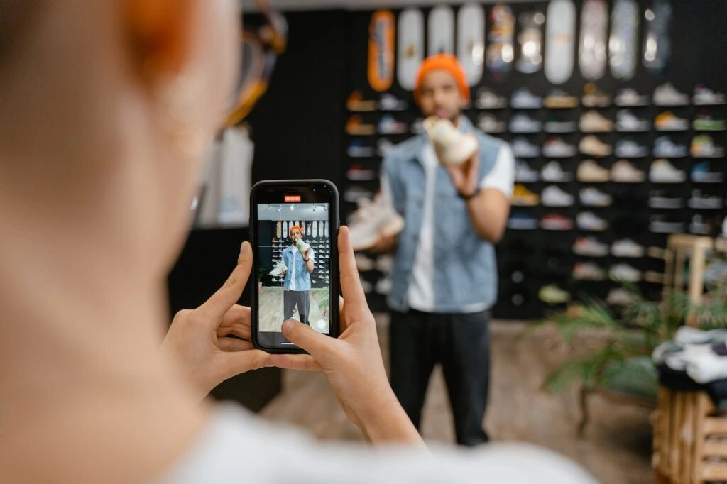 A person records a man showcasing a pair of sneakers during a live stream in a shoe store.