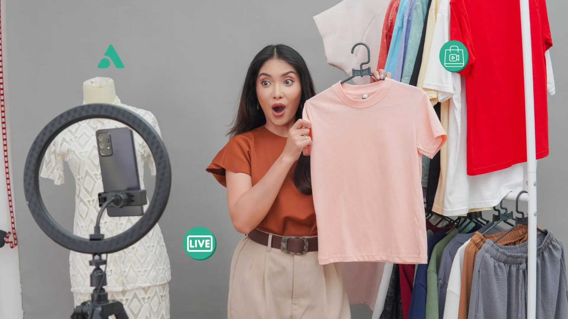 Seorang hos wanita sedang melakukan sesi live selling pakaian menggunakan lampu ring light dan telefon pintar untuk TikTok dan Shopee Live.